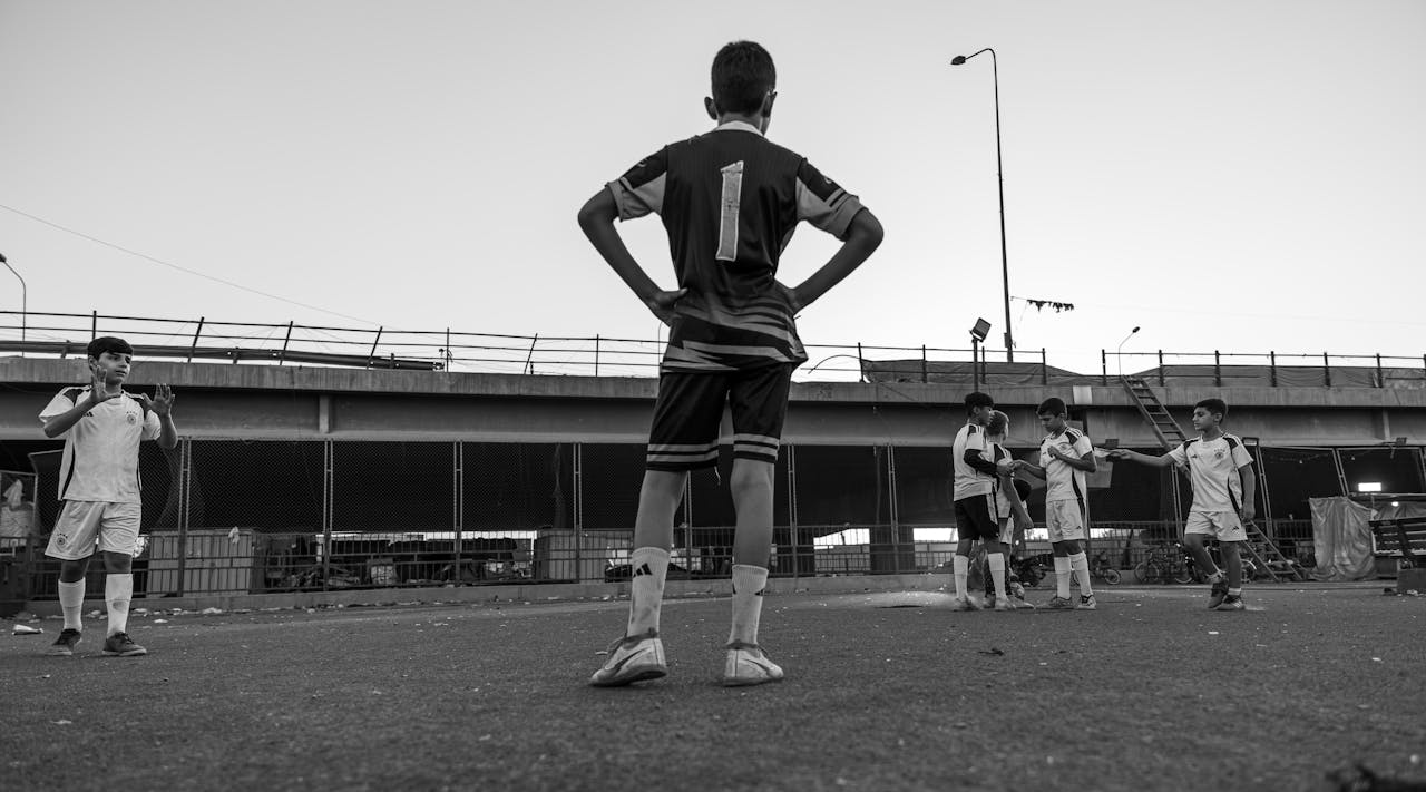 Black and white photo of boys playing soccer in an urban setting under a bridge.